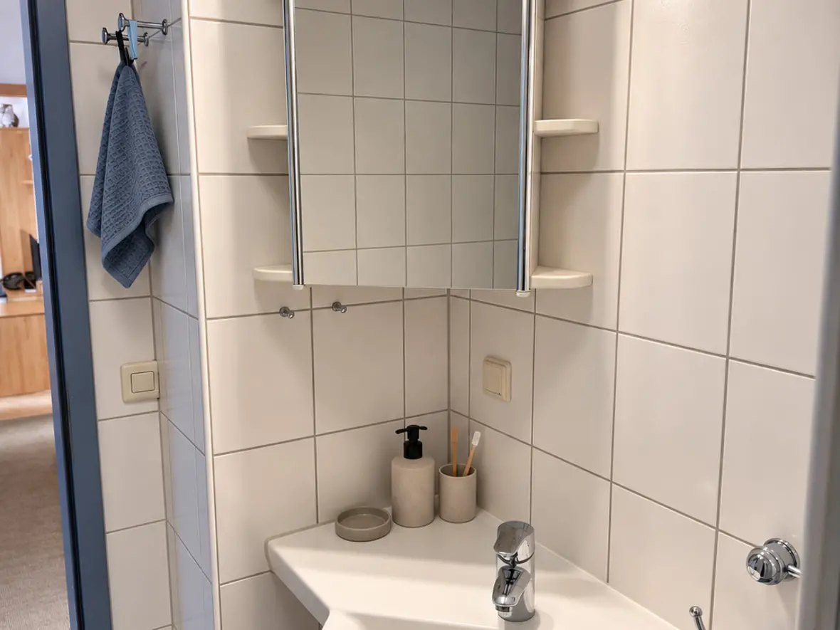 Weißes Eck-Waschbecken mit Einhebelmischer unter einem beleuchteten Spiegelschrank mit Ablagen, daneben blaue Handtücher.