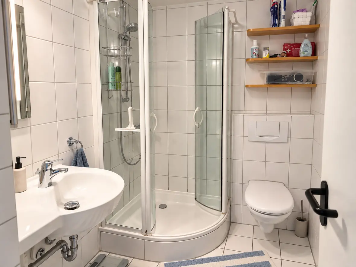 Weiß gefliestes Badezimmer mit runder Eckdusche, Hänge-WC, Waschbecken und Holzregalen an der Wand.