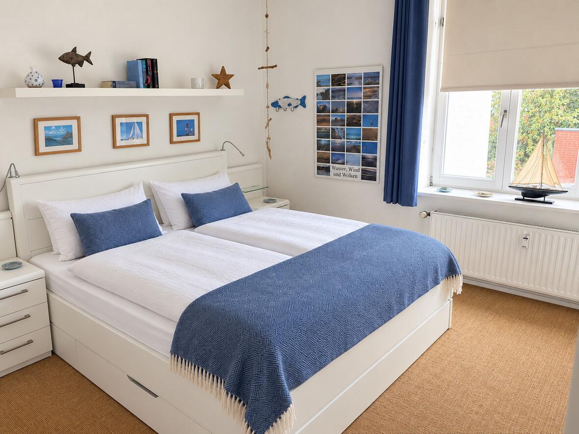 Helles Schlafzimmer mit weißem Doppelbett, blauer Tagesdecke, maritimen Bildern und einem Segelboot-Modell am Fenster.