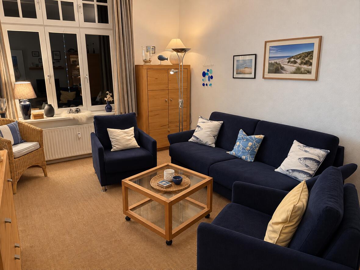 Gemütlich beleuchtetes Wohnzimmer mit dunkelblauer Sofagruppe, Fischkissen, Korbsessel und einem Dünenbild an der Wand.