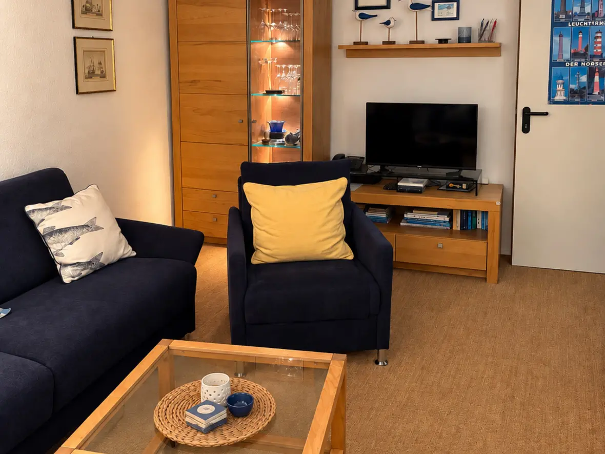 Gemütliches Wohnzimmer im warmen Abendlicht mit dunkelblauem Sofa und Sessel, Couchtisch aus Holz und Glas, beleuchteter Vitrine und maritimer Deko.